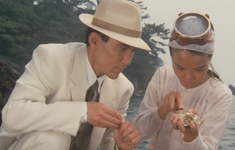 A man crouches next to a young girl shucking an oyster.