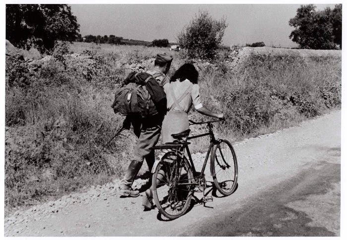 [Italian soldier and woman with bike following captured comrades as they march off to a POW camp, near Nicosia, Sicily]