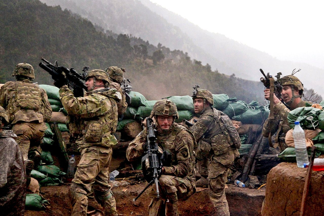 Seven American soldiers in beige tactical gear huddle behind a row of green sandbags on a mountainside, pointing rifles in various directions.