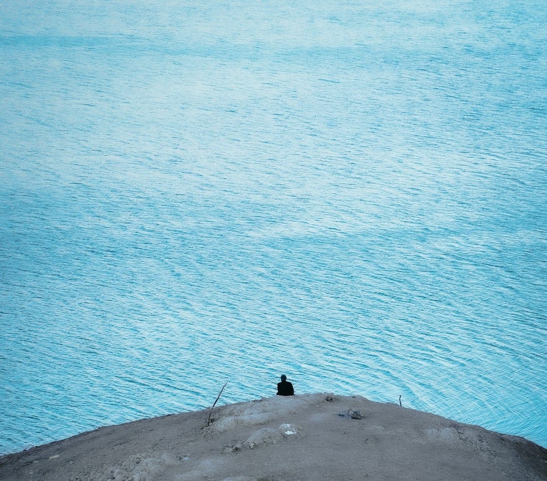 A lone figure sits on a hill overlooking the ocean.