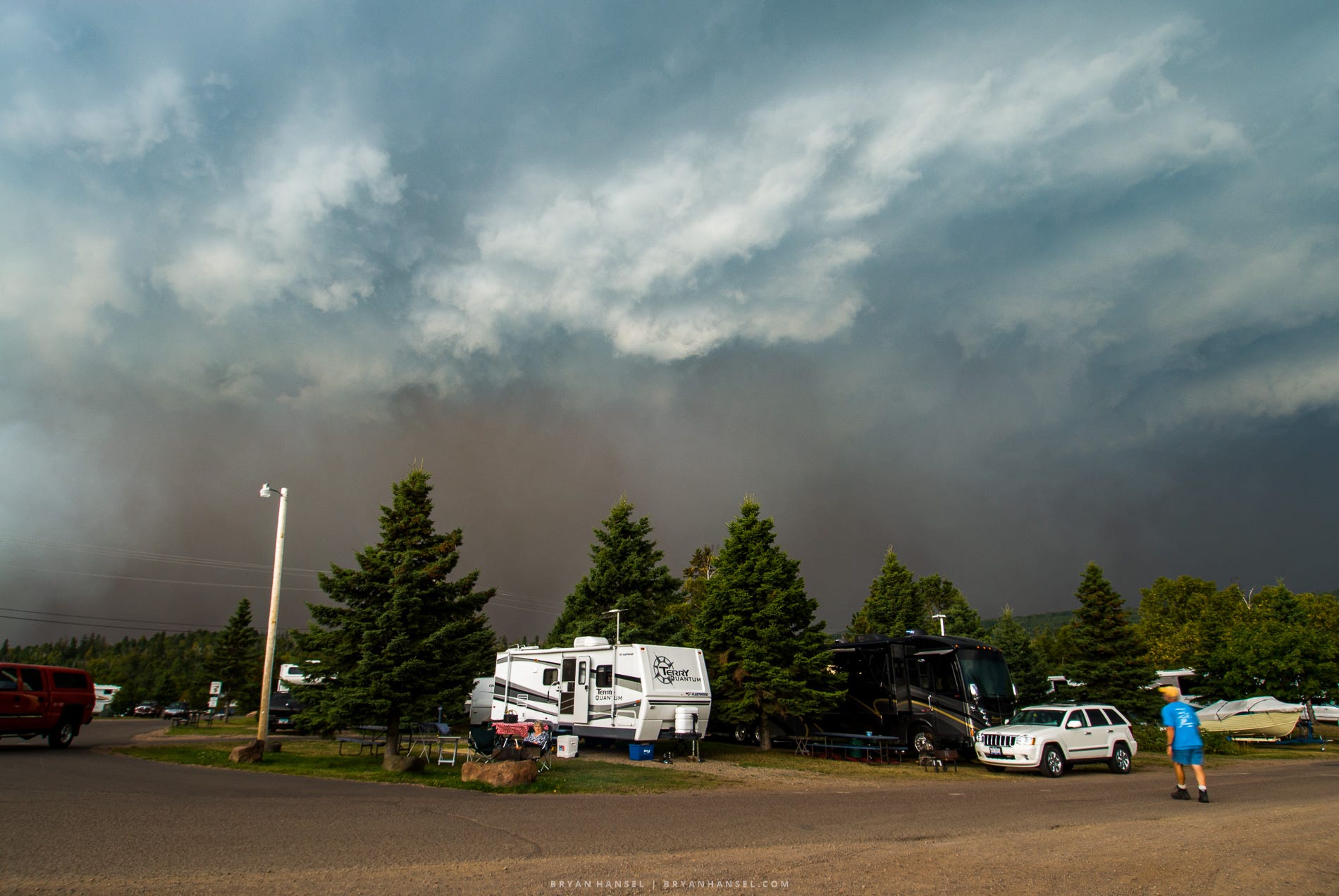 A campground near Grand Marais sits under towering storm clouds and a dark wall of wildfire smoke rising behind the trees as people continue their evening routines.