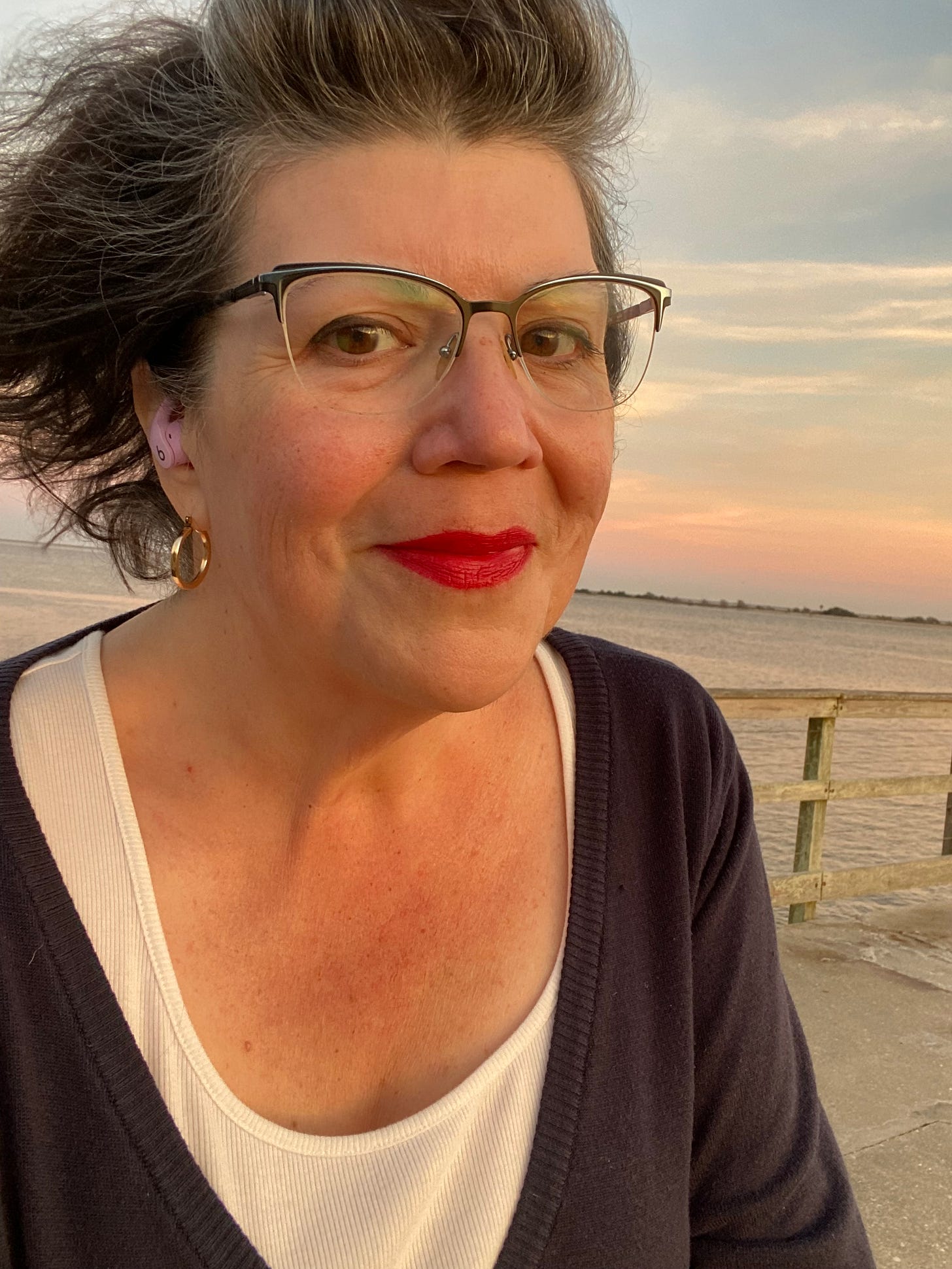 a head/shoulders selfie of a middle aged white woman with greying dark hair, eyeglasses, and bright lipstick. She is wearing a white scoop neck t-shirt under a navy blue cardigan, gold hoop earrings, and lavender earbuds in her ears. Behind her you can see a body of water and a wooden pier railing. The photo is taken during sunset. a head/shoulders selfie of a middle aged white woman with greying dark hair, eyeglasses, and bright lipstick. She is wearing a white scoop neck t-shirt under a navy blue cardigan, gold hoop earrings, and lavender earbuds in her ears. Behind her you can see a body of water and a wooden pier railing. The photo is taken during sunset.
