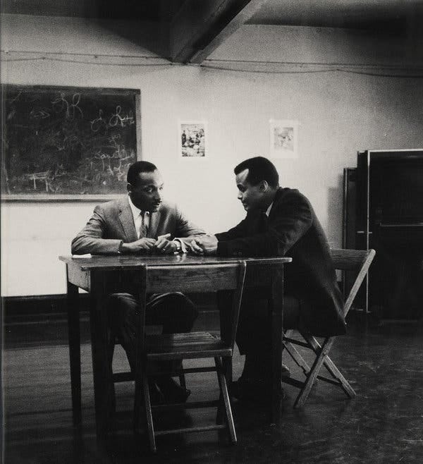 A black and white photo of Mr. Belafonte, in a dark suit and tie, sitting at a small table in a bare room with Martin Luther King, who wears a gray suit and tie. They sit on wooden folding chairs. A black and white photo of Mr. Belafonte, in a dark suit and tie, sitting at a small table in a bare room with Martin Luther King, who wears a gray suit and tie. They sit on wooden folding chairs.