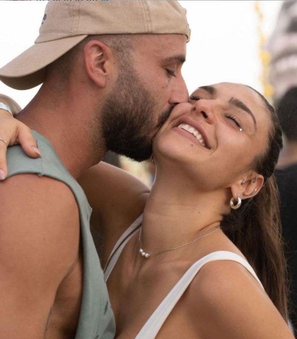 A man wearing a beige cap and green sleeveless top embraces and kisses a smiling woman with long dark hair wearing a white top and pearl necklace and earrings, both appearing joyful in an outdoor setting with blurred background elements like trees and structures.