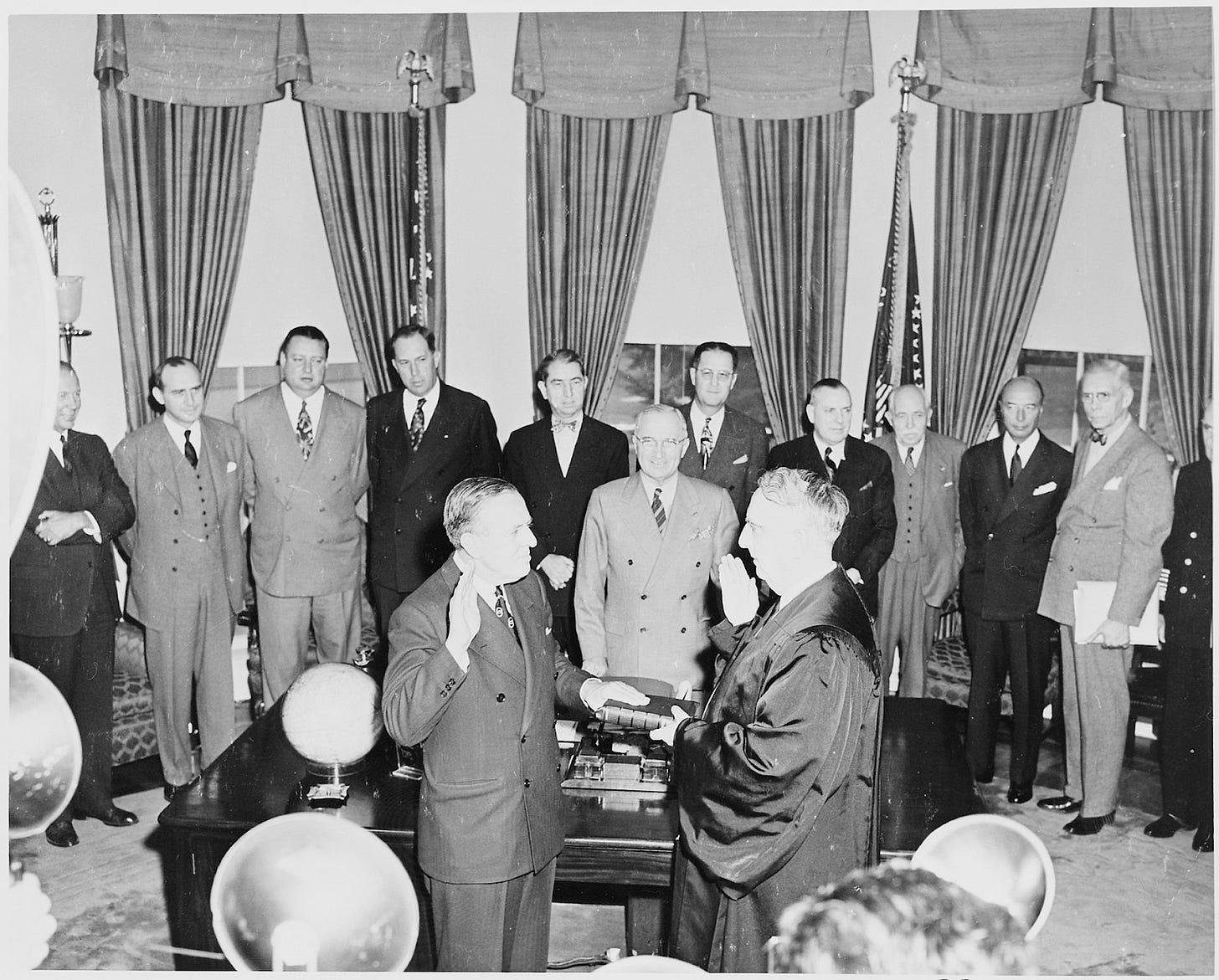 a man in a suit is sworn in by a judge, standing in front of the President's desk in the oval office, with President Truman surrounded by more men in suits.