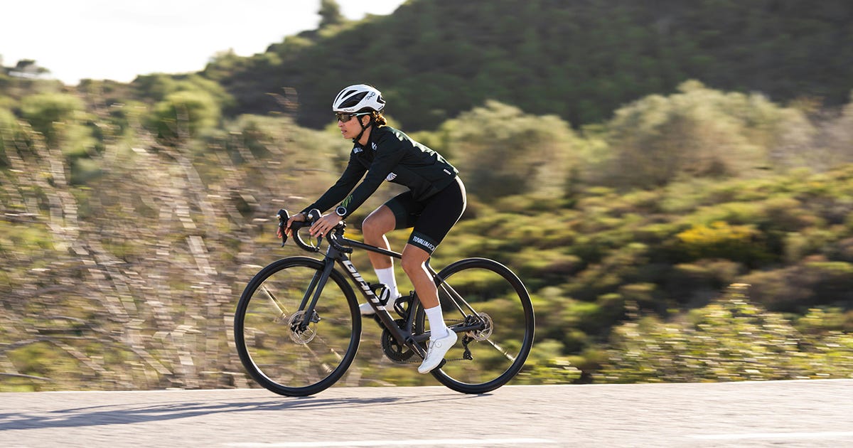 Uma ciclista com roupa preta, capacete e calçados brancos pedala em uma estrada com vegetação seca
