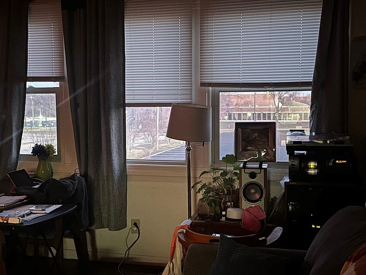 L to r: a photograph of a 100-disc CD changer stacked  and surrounded by newspapers, records, and other ephemera. the lighting of the photo is blue; a corner of a living room in which a lamp stands. 3 windows with the blinds raised can be seen. a small table with speakers and plants is also in the room.