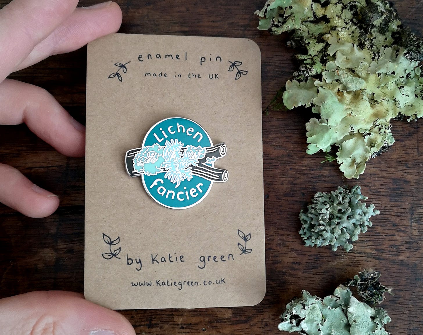 A white person's hand holds and enamel pin on a card. The pin says Lichen Fancier in silver text, and shows a twig adorned with various bluegreen lichens. There is also a selection of real lichens on the wooden table beside it A white person's hand holds and enamel pin on a card. The pin says Lichen Fancier in silver text, and shows a twig adorned with various bluegreen lichens. There is also a selection of real lichens on the wooden table beside it
