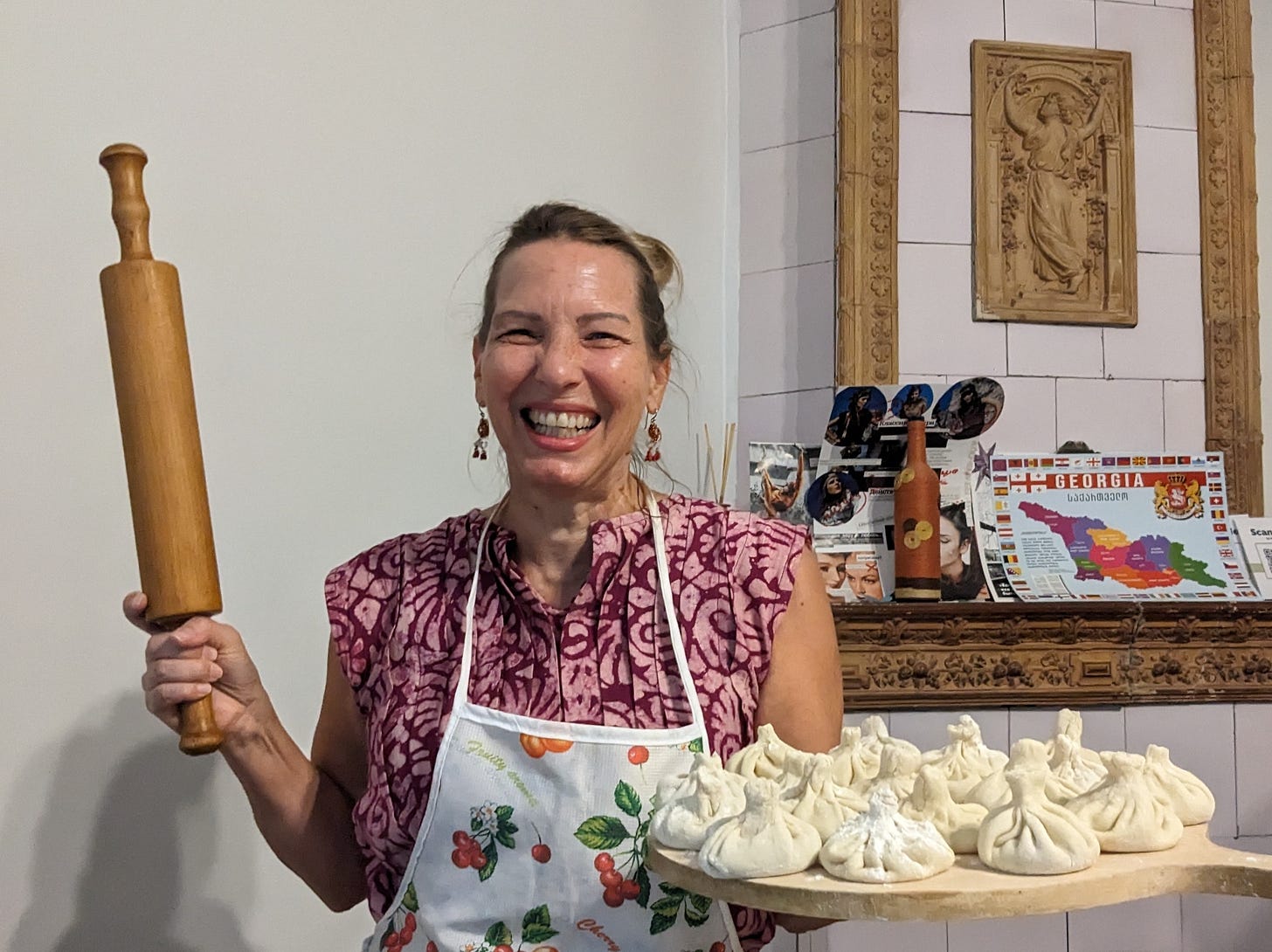 A white woman is wearing a pink blouse and is holding rolling pin and a tray of uncooked pastries.  She is inside someone’s living room and laughing. 