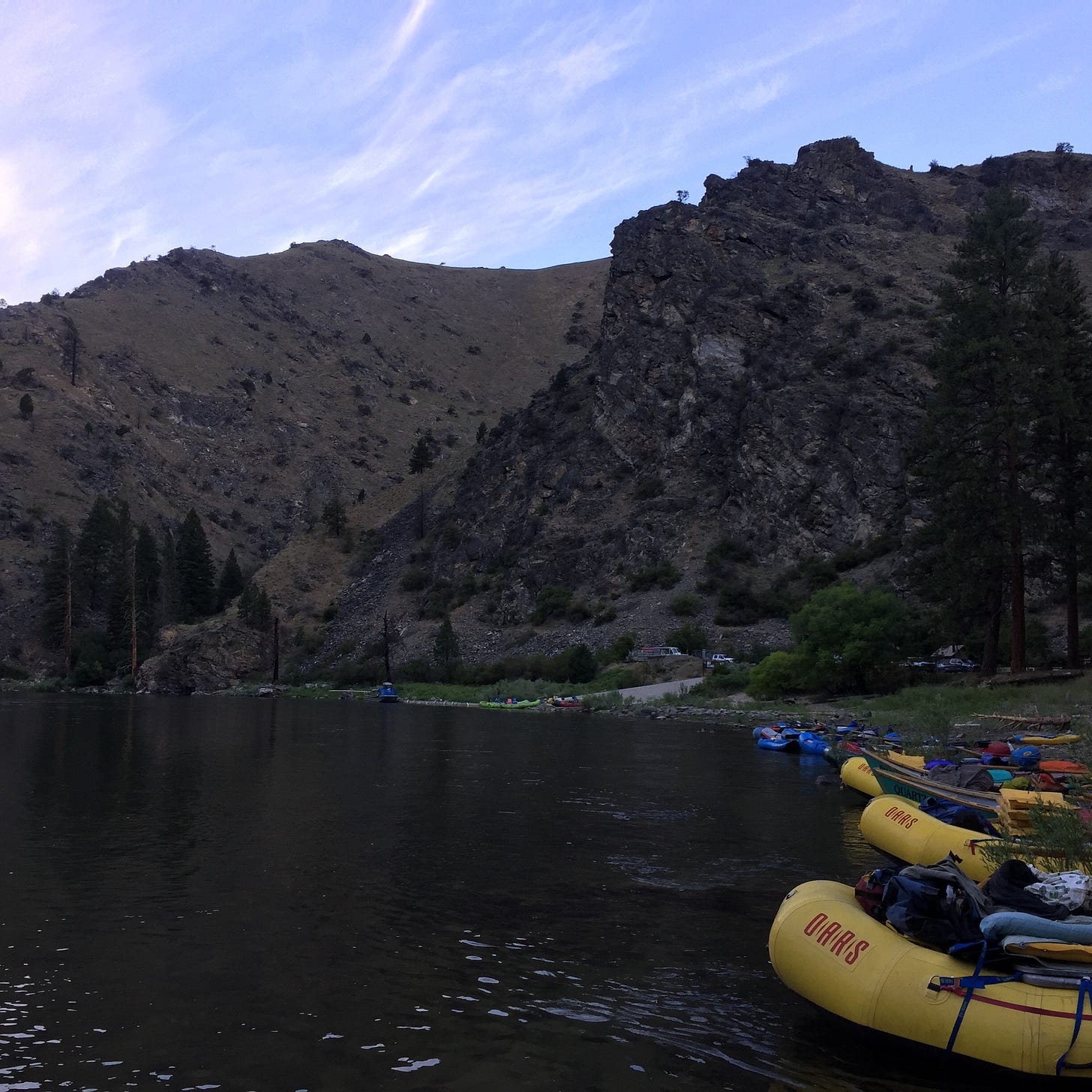 Looking downstream at a river canyon as the sun sets. There are craggy grey and green mountains, an inky blue river, and at least ten rubber boats of assorted colors.
