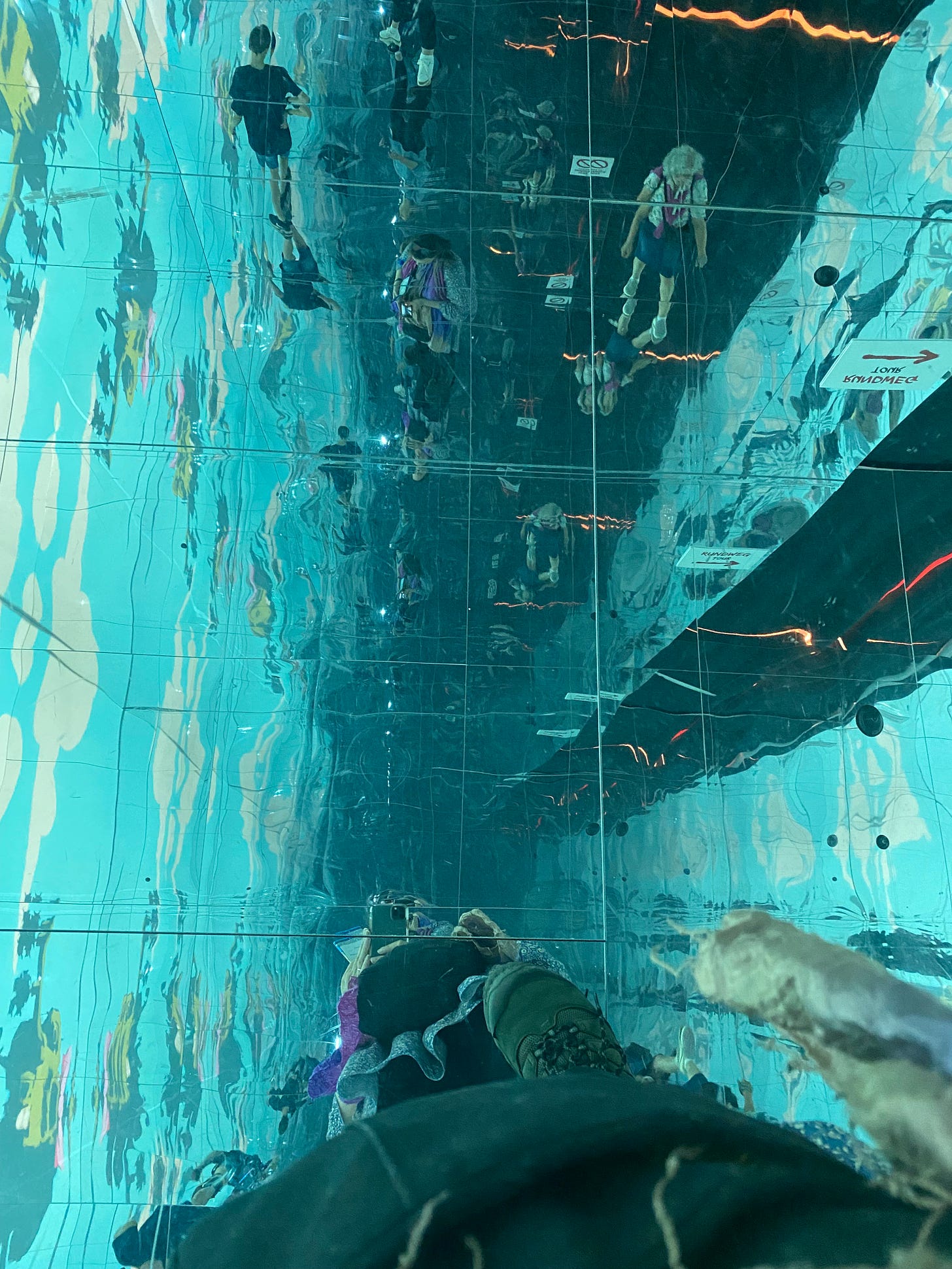 a turquoise field of reflections in an infinity room with a closeup of boots at the base of the image and a reflection of the phone camera above them