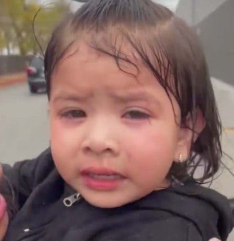 Close-up photograph of a young child with wet curly hair wearing small earrings and a black hooded jacket, sitting in a car seat with a distressed expression, eyes squinted and mouth slightly open, against a blurred background of a parking lot with vehicles and buildings.