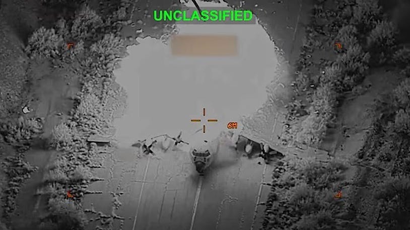 aerial black and white video screenshot of a parked cargo plane exploding in a fireball. A red targeting reticle and the word "unclassified" in bright green letters are superimposed over the image. 