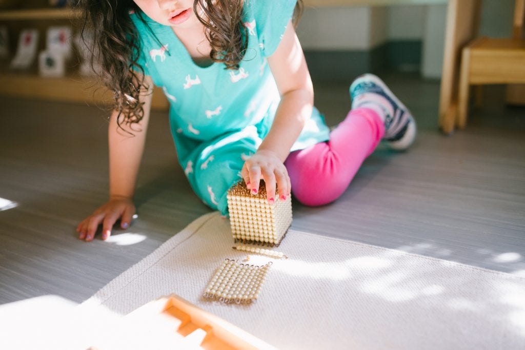 Young girl kneeling on the floor of a Montessori classroom, working independently with golden bead math material