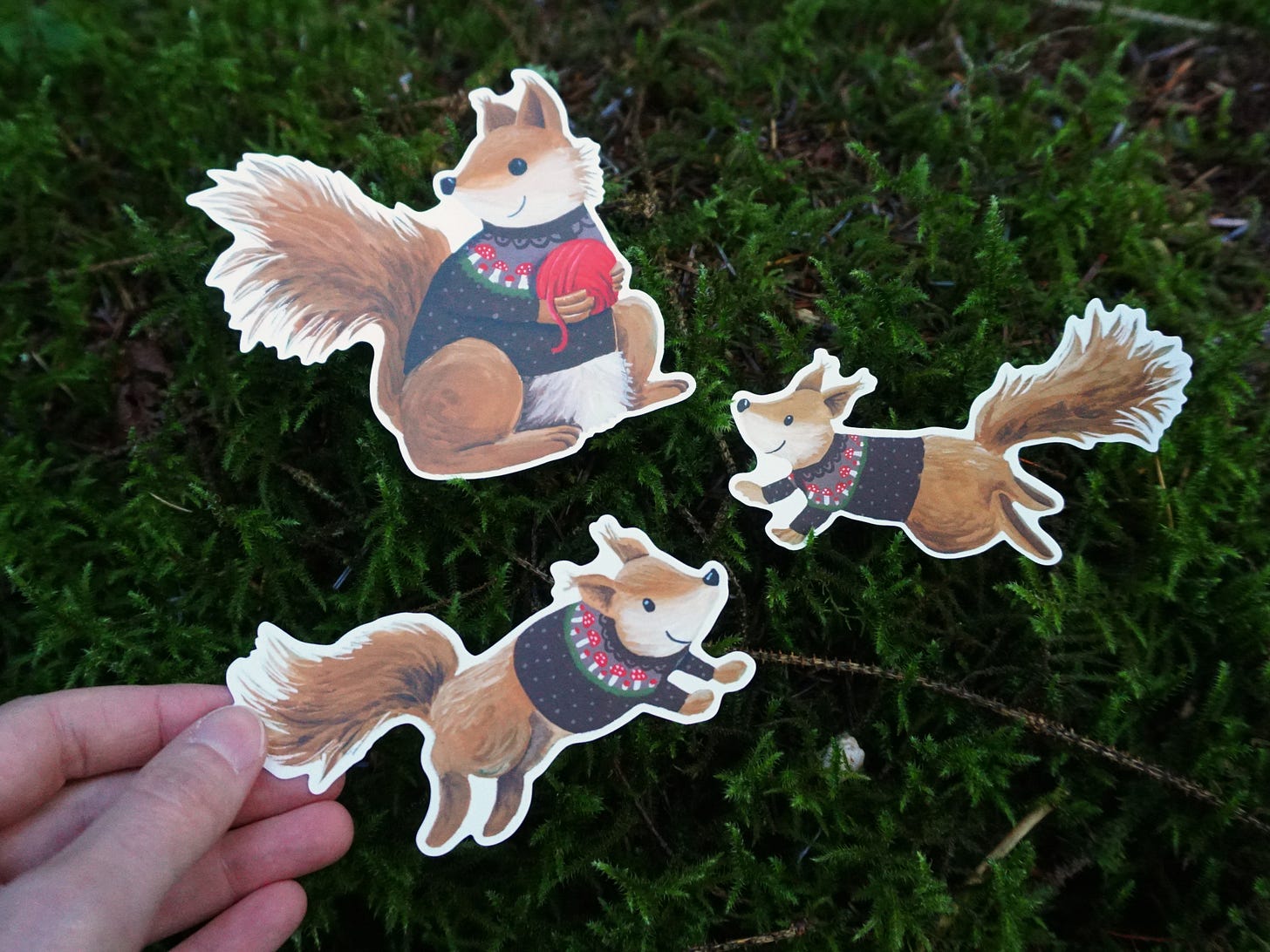 Image description: three shaped sickers of red squirrels wearing mushroom cardigans. The largest on is holding a ball of red yarn. The smaller two are bouding with joy.