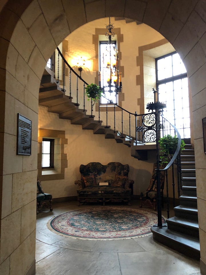 Four views of a circular stair well. From top-left to bottom-right: view of a circular carpet and two-seater sofa below a wrought-iron staircase with windows; a sidetable on which a suit of armour rests, with a carpet showing a unicorn on the wall and a tall-backed chair near a window; a view up the staircase showing wrought-iron light fittings; windows, a curving ironwork staircase, and hanging ironwork light fittings.