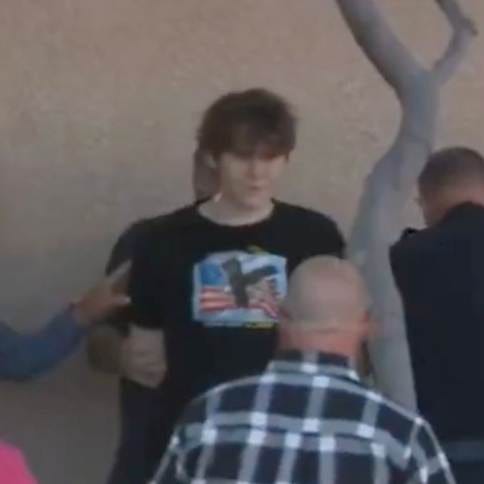 Tyler Robinson wearing a black long-sleeve shirt with an eagle and American flag design, standing outdoors near a tree and a concrete wall. Multiple people, including a man in a plaid shirt, are visible around him. The shirt design includes the text "Land of the Free Home of the Brave."