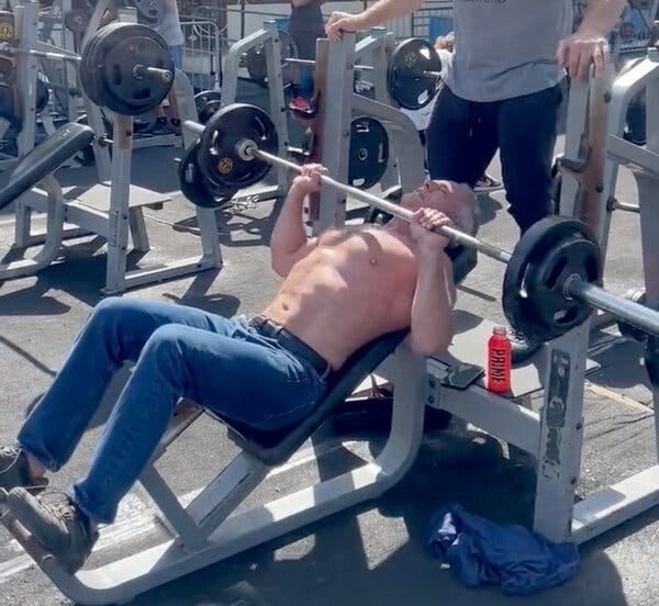 Robert F. Kennedy Jr., shirtless and wearing jeans, lifts weights at an outdoor gym with a large man standing behind him spotting him. 