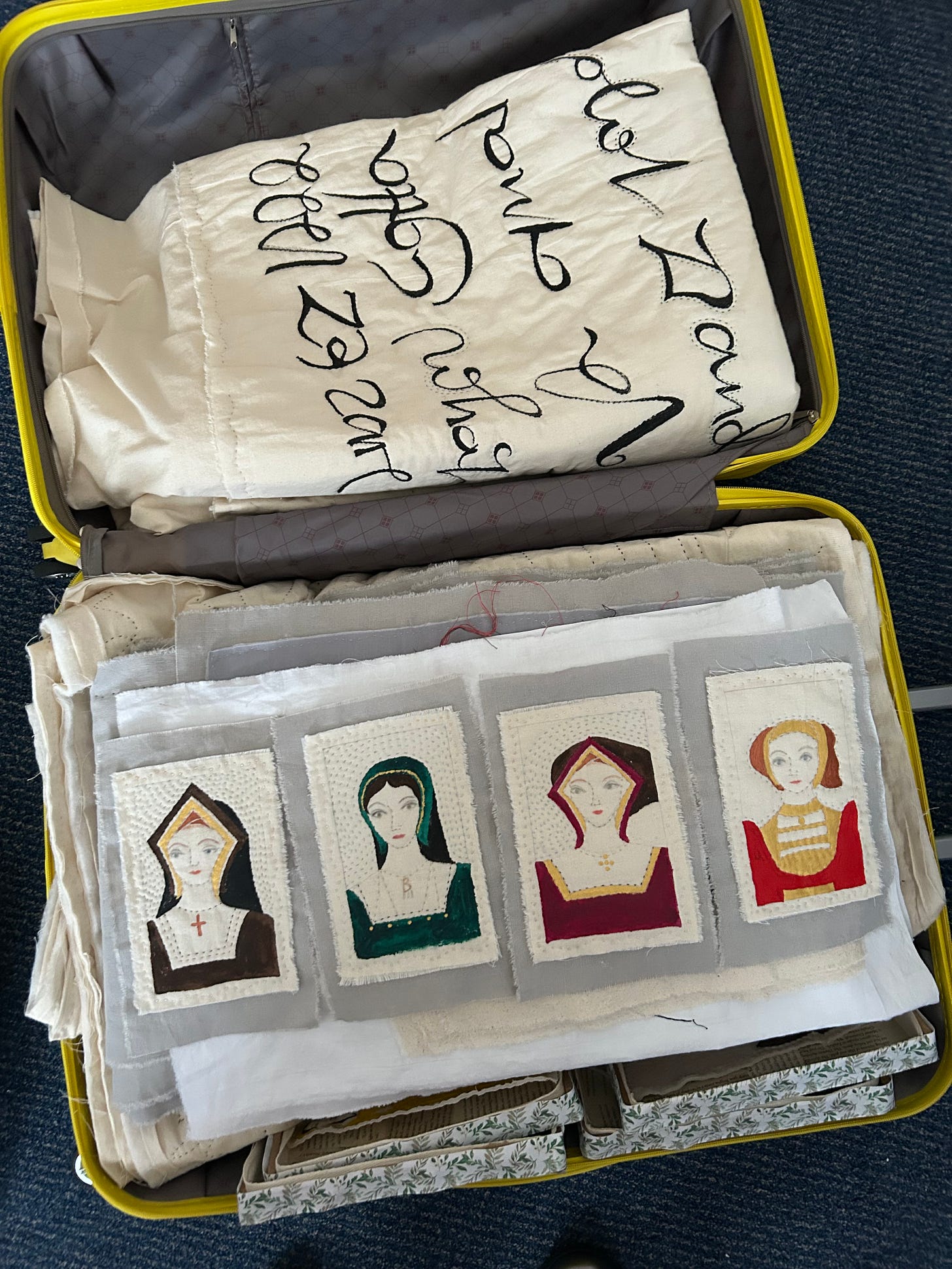 An open yellow suitcase containing textile pieces, at the back is a folded piece with 16th century lettering, at the front four small portraits of Katherine of Aragon, Anne Boleyn, Jane Seymour and Anna of Cleves - painted on fabric and stitched.