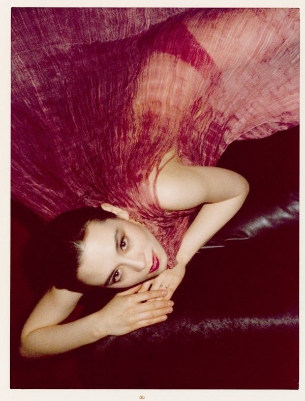 A short-haired, fair woman wearing a deep pink pleated silk dress rests her head on her folded hands and looks at the camera.