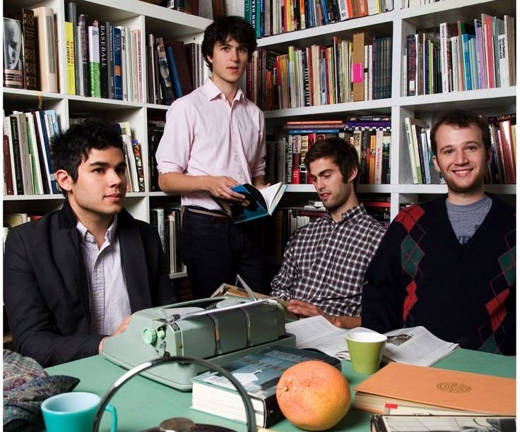 This may contain: three young men sitting at a table in front of bookshelves and an orange