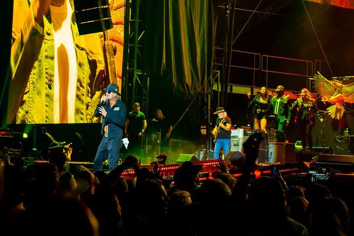 Kid rock is wearing a black open face button down, black hat and black sun glasses singing on stage with his band while the crowd dances to his music
