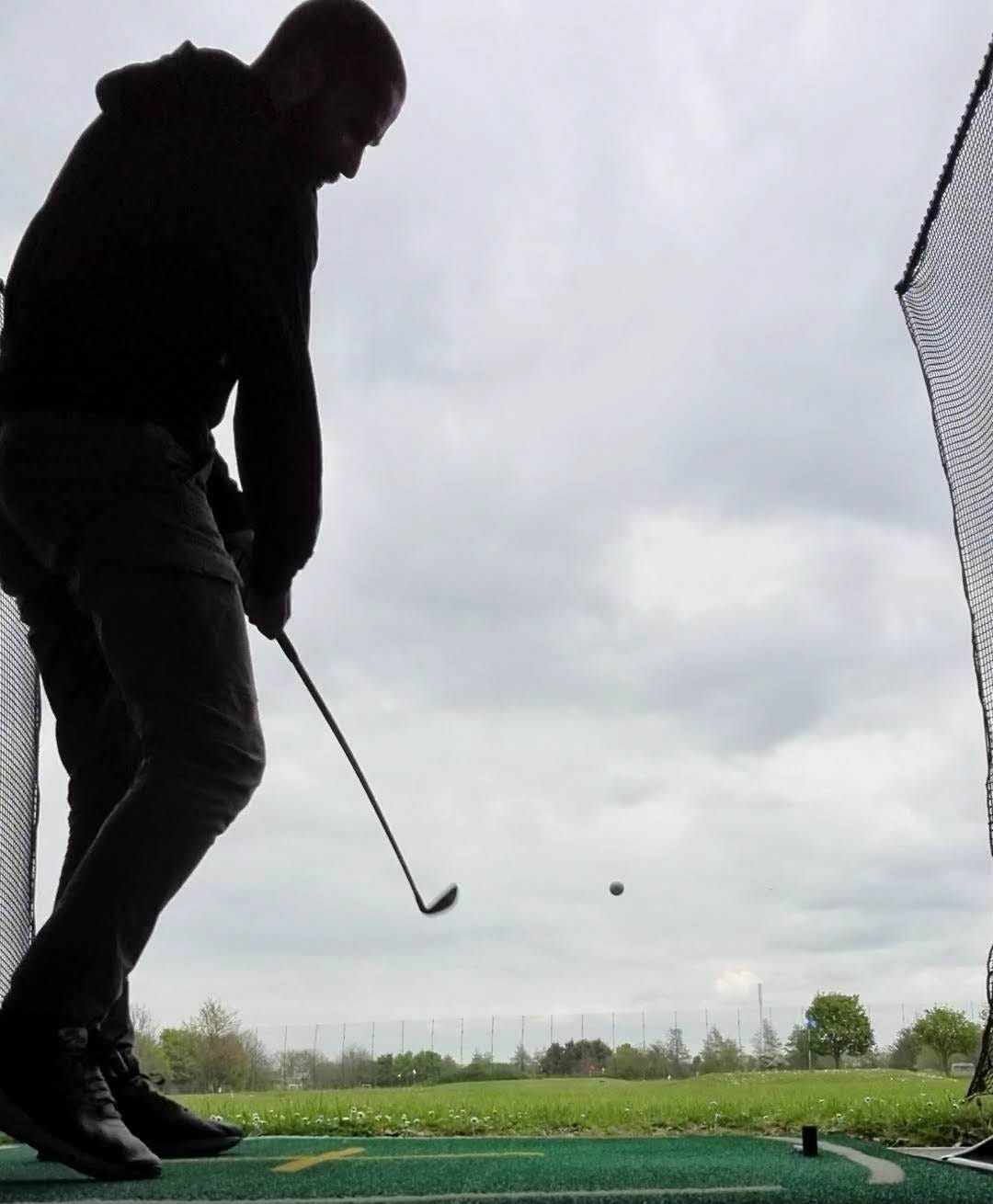 Paul hitting a golf ball at an outdoor driving range, silhouetted against a cloudy sky with the ball visible in mid-air.
