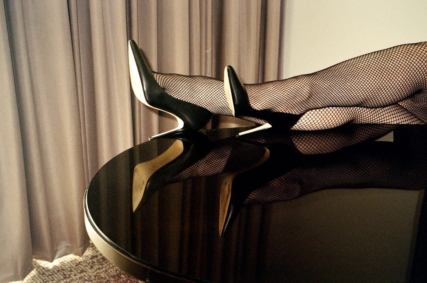 Evyn Silver's feet with the ankles crossed, resting on a reflective dark wood desk. She's wearing black fishnets and black leather pumps. In the background is a light taupe curtain and a white wall. Evyn Silver's feet with the ankles crossed, resting on a reflective dark wood desk. She's wearing black fishnets and black leather pumps. In the background is a light taupe curtain and a white wall.