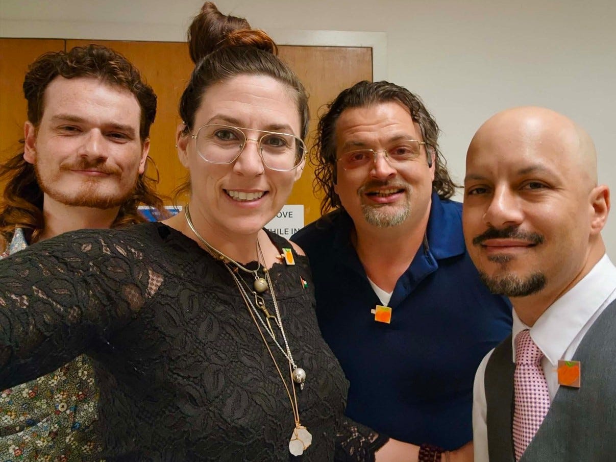 Four people pose for a selfie in a courthouse hallway, smiling at the camera. All four are wearing small orange peach pins. The person taking the photo is wearing a black lace top and glasses. The others wear a floral shirt, a blue polo, and a suit with a pink tie. Four people pose for a selfie in a courthouse hallway, smiling at the camera. All four are wearing small orange peach pins. The person taking the photo is wearing a black lace top and glasses. The others wear a floral shirt, a blue polo, and a suit with a pink tie.