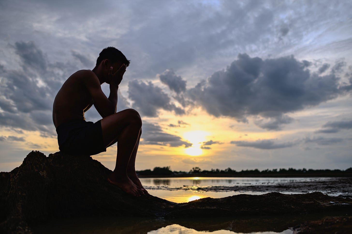 Silhouette image of young man sitting with hands over face. Sunset in background. Silhouette image of young man sitting with hands over face. Sunset in background.