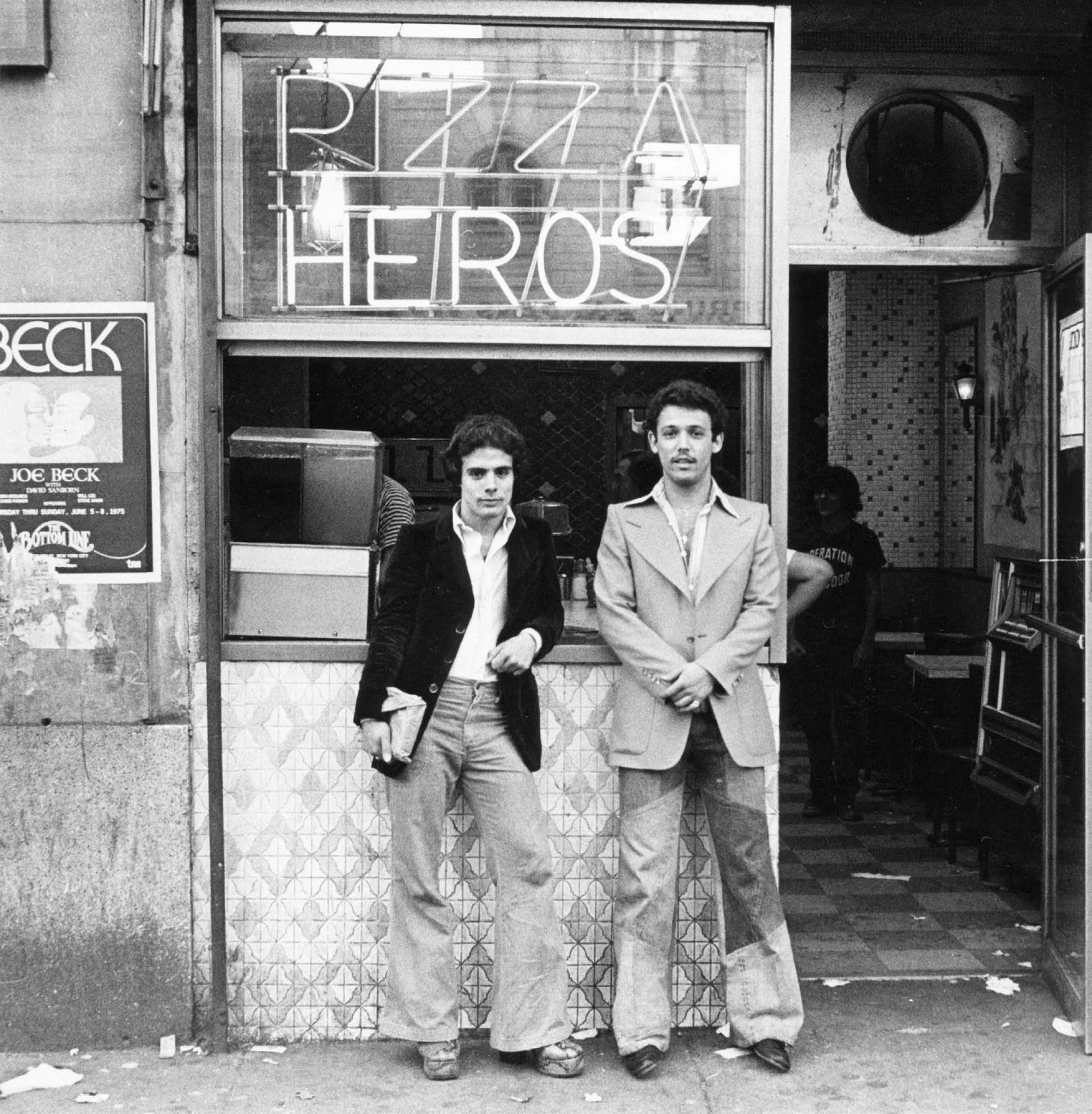 Two men stand in front of a storefront with Pizza Heros sign