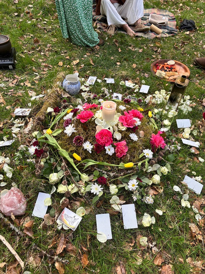L-R: two white women sit in a garden, smiling at the camera, it's very sunny, there's food on the table between them, a mandala on the forest floor with a candle, flowers and cards, trees in the forest, three crocuses, two white, one purple
