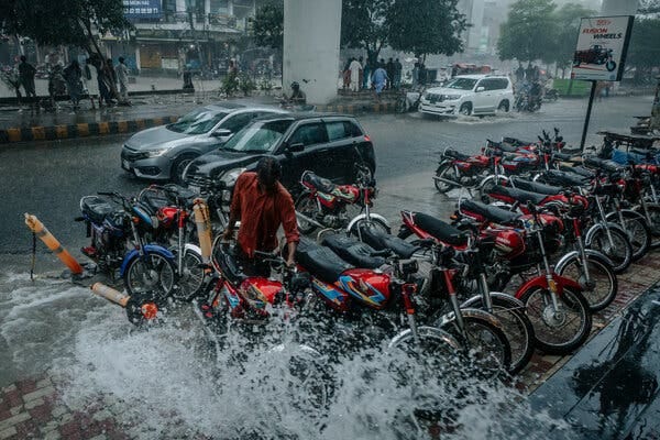 A person parks a motorcycle alongside a couple dozen others in a heavy rainstorm as water splashes over the bikes.