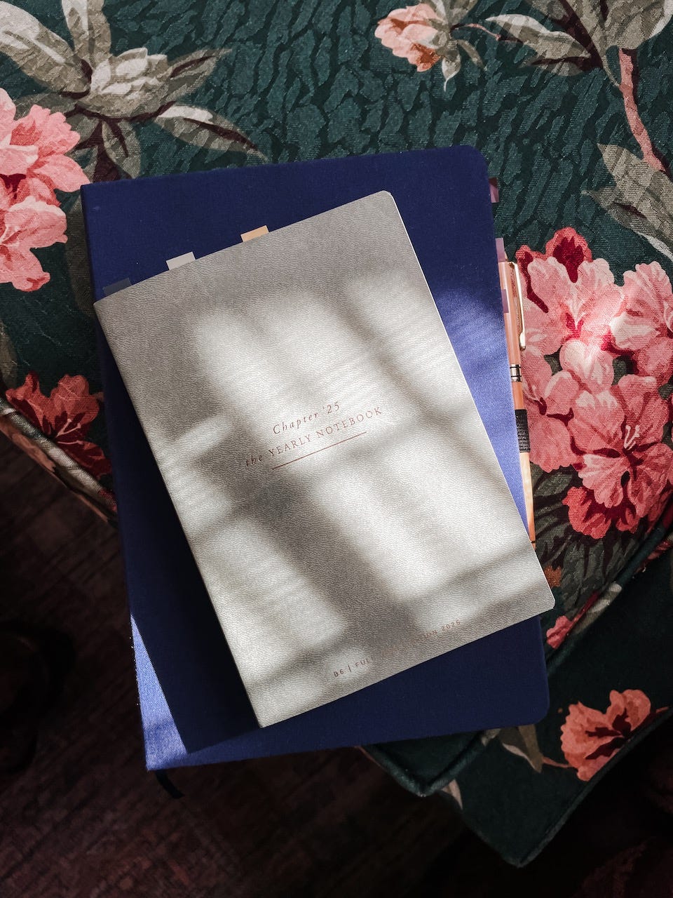 An agenda with "chapter '25" sits on another notebook. Both journals rest on a flowery patterned sofa cushion.