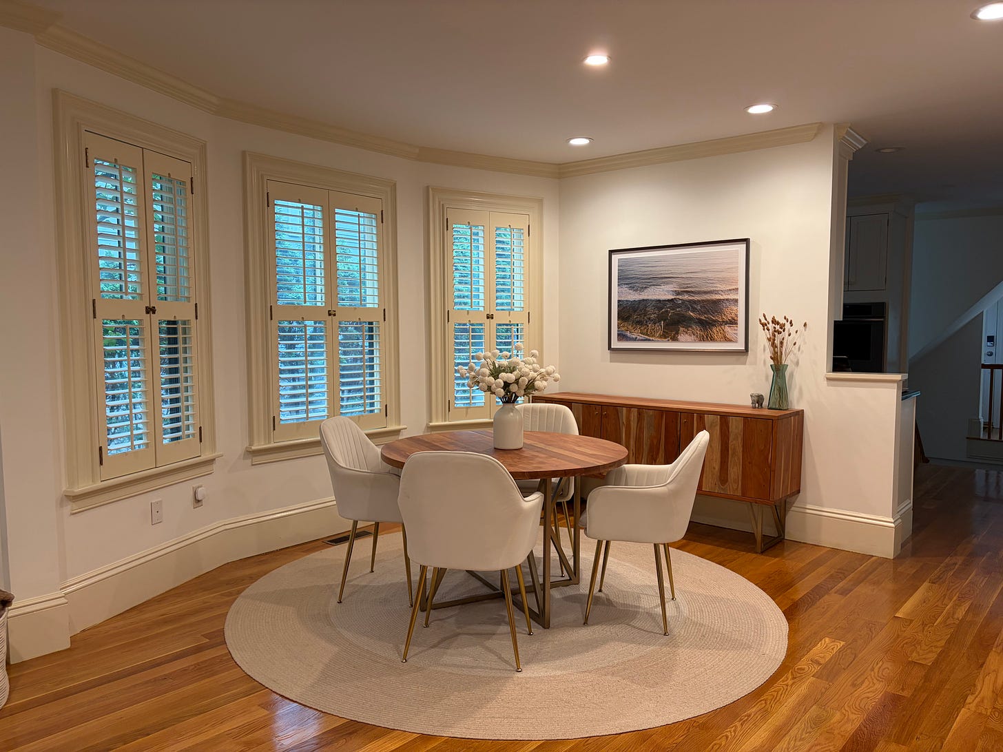 Dining room in a classic colonial home with a wood table, black chairs, bay window, and a framed ocean photograph above a dark wood sideboard.