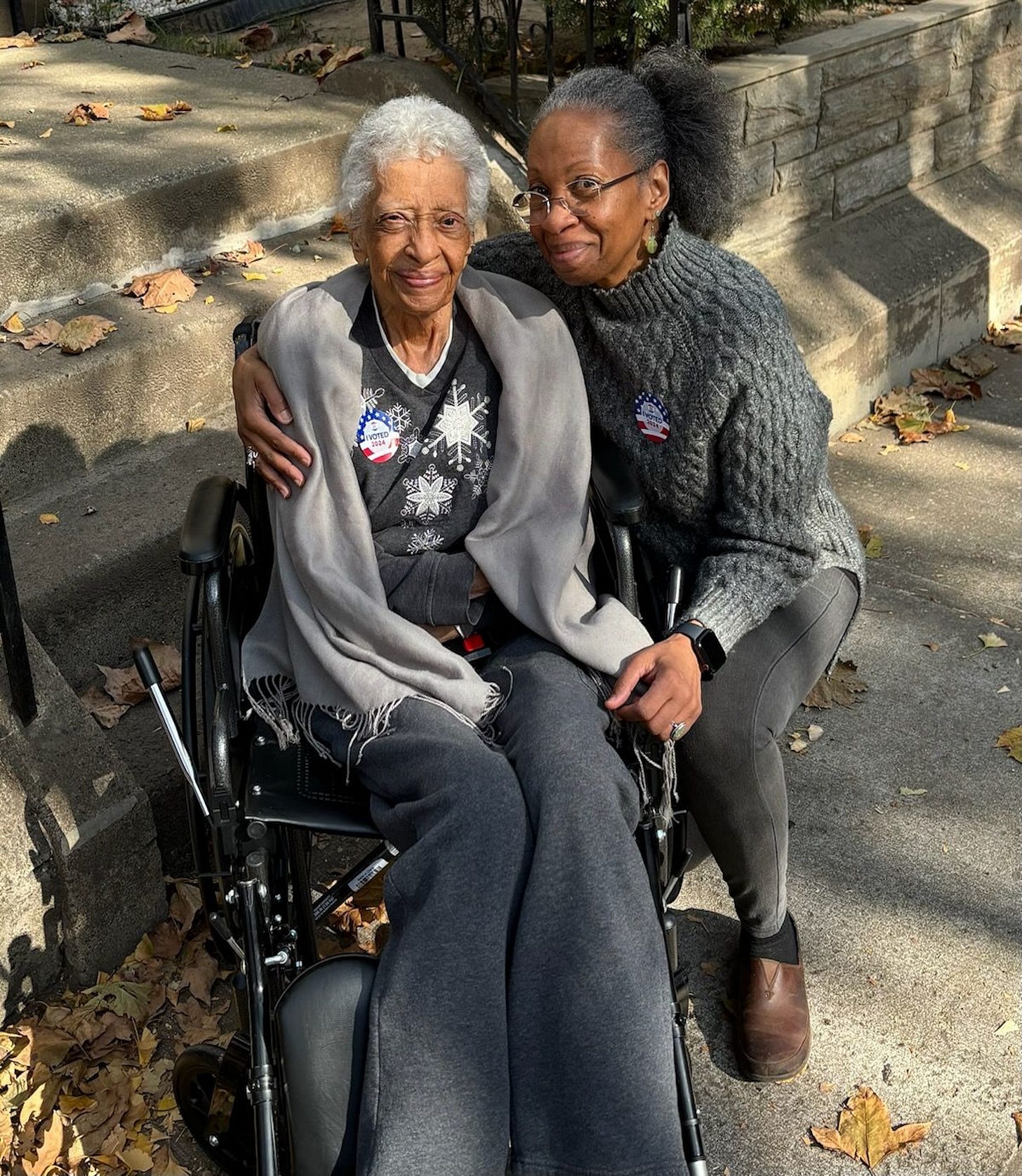 A portrait of two women side-by-side. On the viewer's left is a senior woman in a wheelchair. On the right is a middle-aged woman who has her arm around the senior woman as she kneels beside her. They are both smiling warmly. A portrait of two women side-by-side. On the viewer's left is a senior woman in a wheelchair. On the right is a middle-aged woman who has her arm around the senior woman as she kneels beside her. They are both smiling warmly.