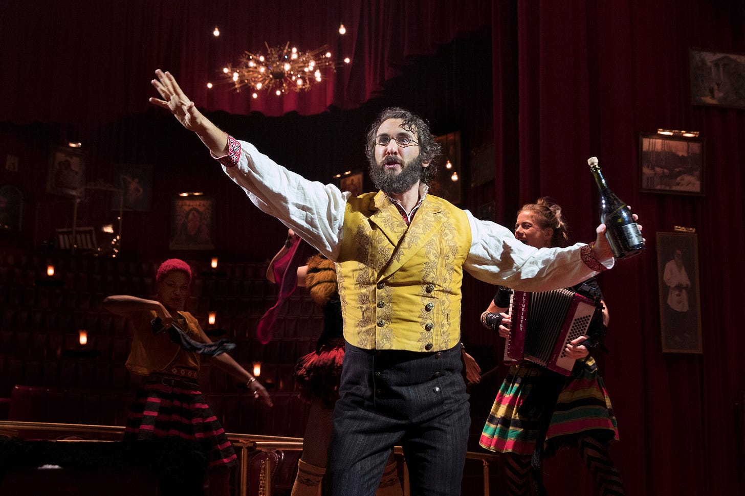 Josh Groban, a dark haired man with a beard and in a yellow waistcoat, with his arms open and holding a bottle of wine
