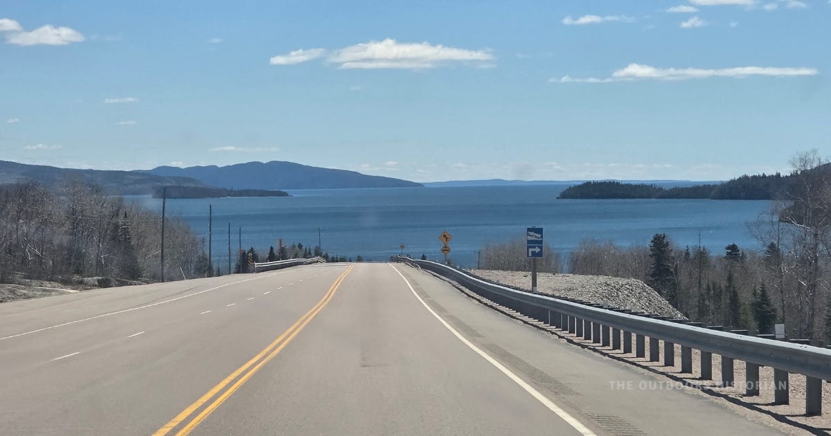 View through the windshield of a vehicle driving along Highway 17 on the eastern shore of Lake Superior. Blue water, forested islands, and a sunny blue sky are visible ahead.