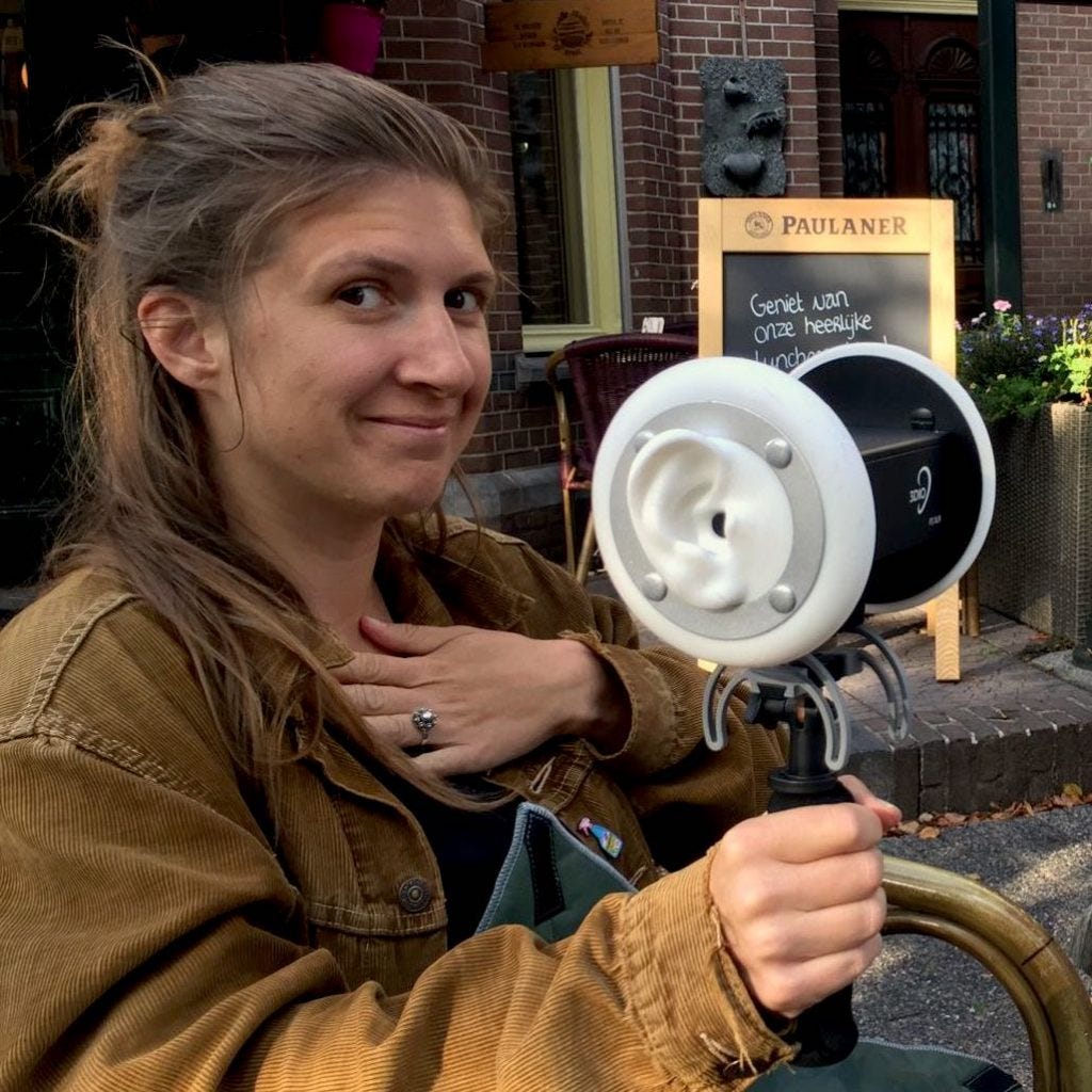 Foto van Jozien (bruin lang haar, bruin vest) buiten met een binaurale microfoon. Dat is een waarbij de positie van de oren nagebootst wordt, het ziet er heel futuristisch uit.