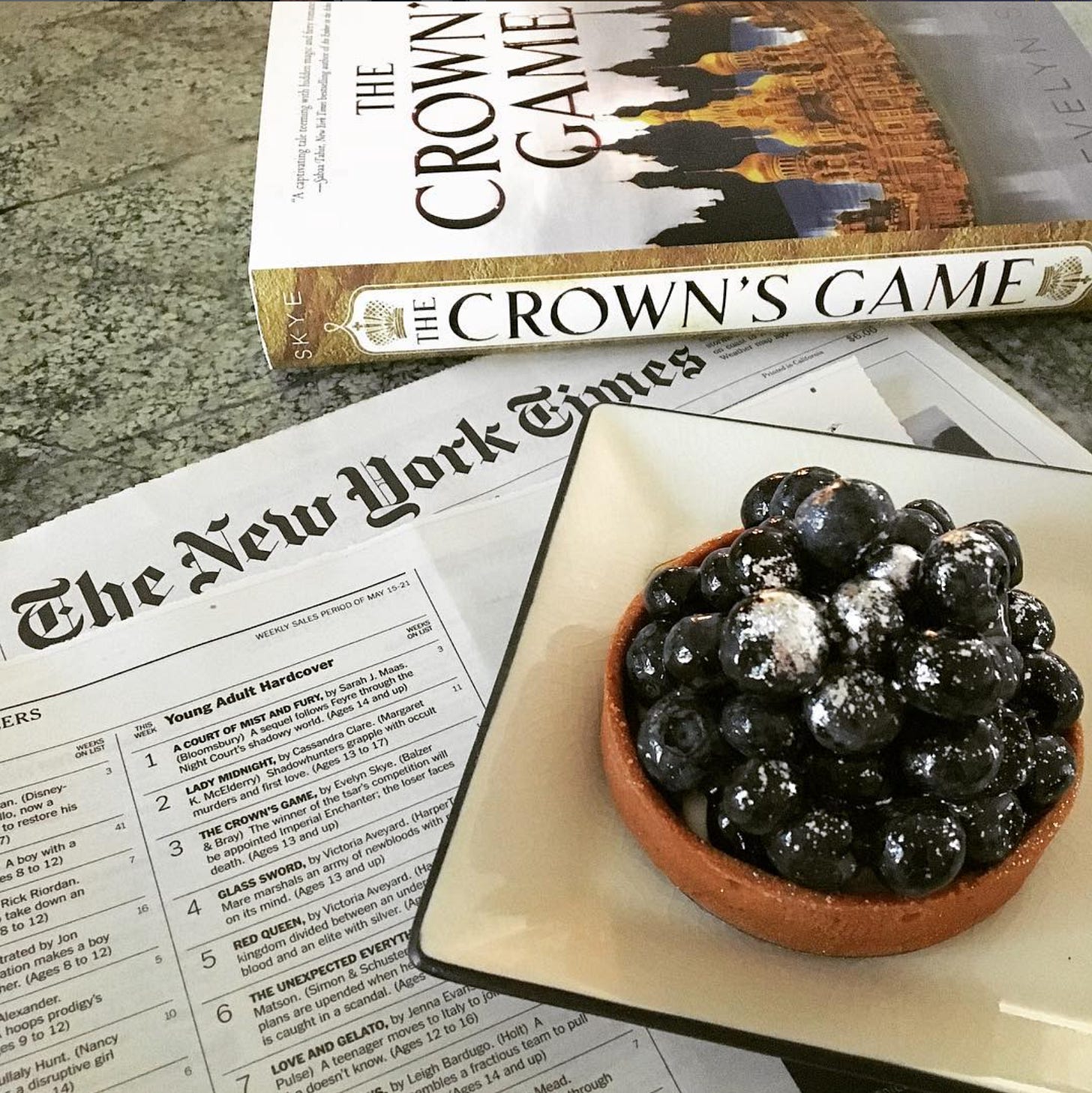 Hardcover copy of The Crown's Game, New York Times newspaper opened to the bestseller list, and a blueberry tart with silver leaf decoration Hardcover copy of The Crown's Game, New York Times newspaper opened to the bestseller list, and a blueberry tart with silver leaf decoration