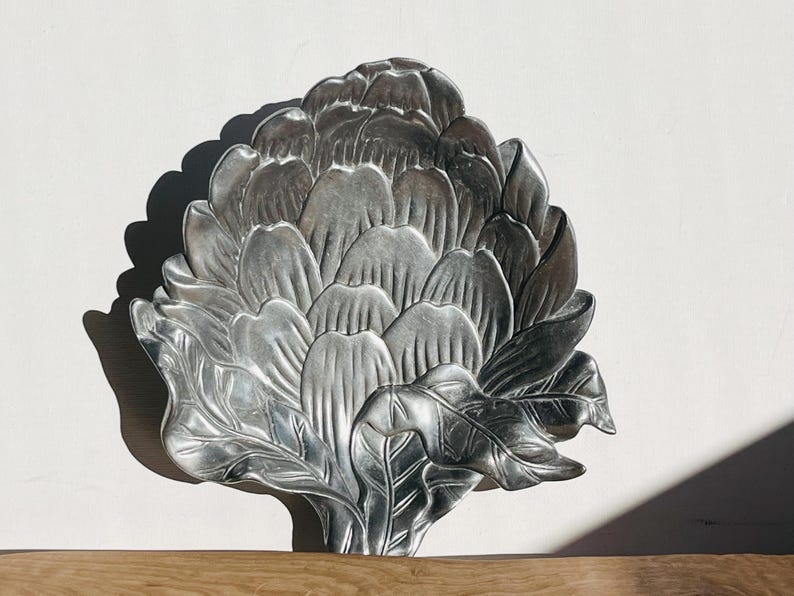 May include: A silver-colored metal artichoke-shaped decorative tray. The tray is intricately designed with detailed artichoke leaves and petals.  It's a unique serving piece or decorative accent for a kitchen or dining setting. The metallic finish gives it a sophisticated look. The piece is shown against a plain white background.