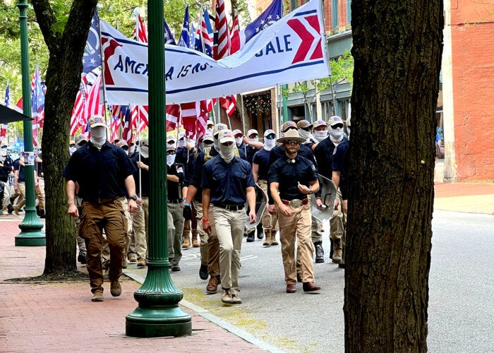 Several white men are shown dressed in navy shirts and khakis. The men wear ball caps and white face masks to hide their faces. They hold upside down American flags and march down a street during the daytime. Several white men are shown dressed in navy shirts and khakis. The men wear ball caps and white face masks to hide their faces. They hold upside down American flags and march down a street during the daytime.