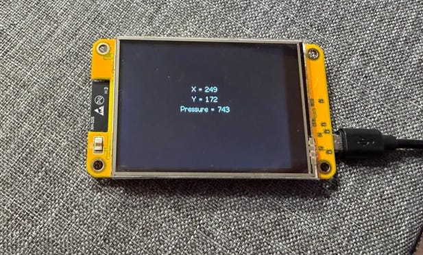 Photo of a yellow ESP32 touchscreen displaying text showing touch coordinates “X = 249, Y = 172, Pressure = 743” on a black background, placed on a gray fabric surface.