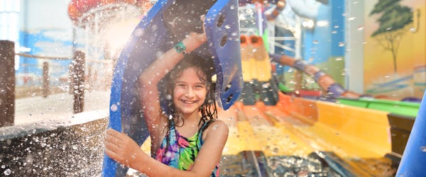 Smiling young girl holding a blue foam mat at the end of a water slide inside Kalahari Round Rock’s indoor water park, surrounded by splashes of water and bright, colorful slide structures in the background.