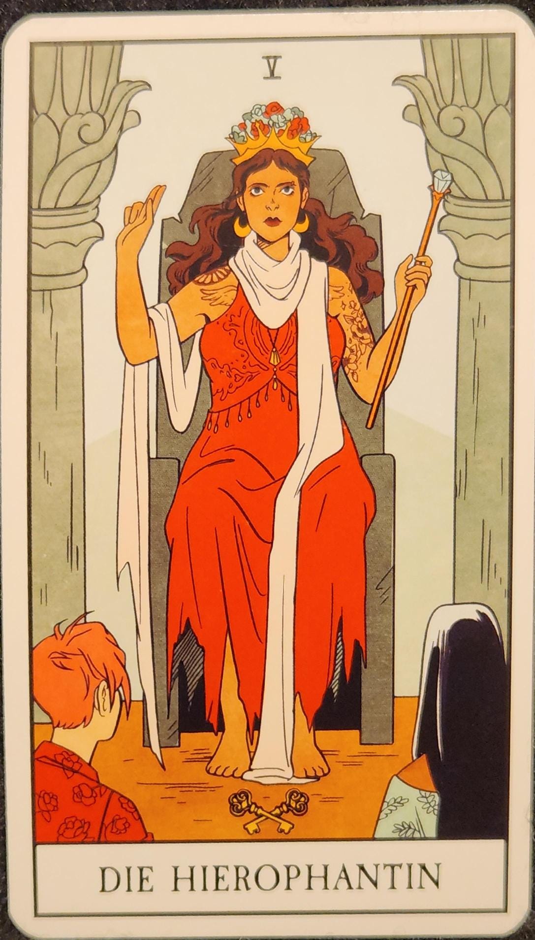 A tarot card labelled V Die Hierophantin. A woman with long wavy brown hair is sitting on a stone throne between two stone pillars. She is wearing a red sleeveless dress with a long white scarf wrapped around her neck and draped over her arm. She is wearing a golden crown with flowers on her head. Her right hand is raised, her fingers held up. In her left hand, she is holding a sceptre or wand. Two keys sit at her feet, crossed over each other. In front of her, two people are kneeling and facing her. 