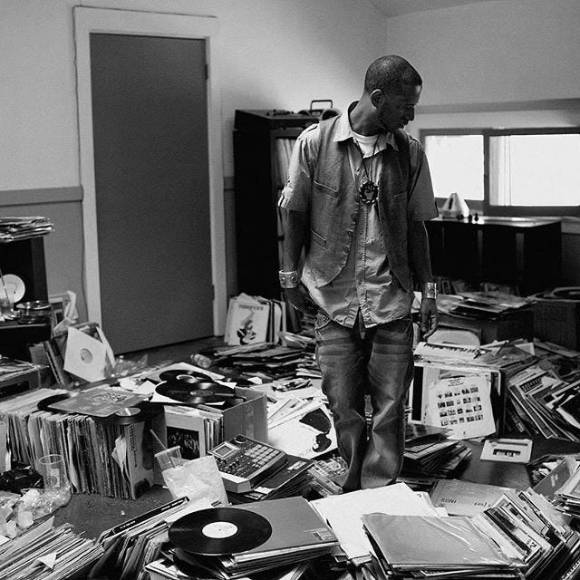Black and white photo of record producer Madlib standing in his studio which is a hot mess of records, some stacked, some spilling in disarray on the floor. He is standing in the looking down as if he searching for something but can't quite remember what. Tragically Madlib's house was burned down in the Los Angeles fire earlier in 2025 and he lost his extensive record collection in the blaze.