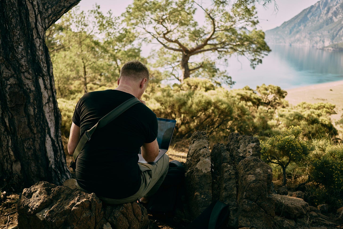 Photo by Anastasia Nelen on Unsplash. Man sitting with a laptop computer under a tree on a hill overlooking a lake. Mountains in the distance. Photo by Anastasia Nelen on Unsplash. Man sitting with a laptop computer under a tree on a hill overlooking a lake. Mountains in the distance.