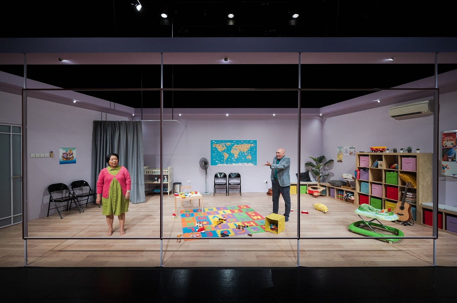 Cat (Neo Swee Lin, left) and David (Lim Kay Siu) in the “cry room” of their church. The room is scattered with toys, there's a map and cot on one side of the room, a series of cubbyholes on the other, and a soft alphabet mat on the floor next to a walker. Cat (Neo Swee Lin, left) and David (Lim Kay Siu) in the “cry room” of their church. The room is scattered with toys, there's a map and cot on one side of the room, a series of cubbyholes on the other, and a soft alphabet mat on the floor next to a walker.