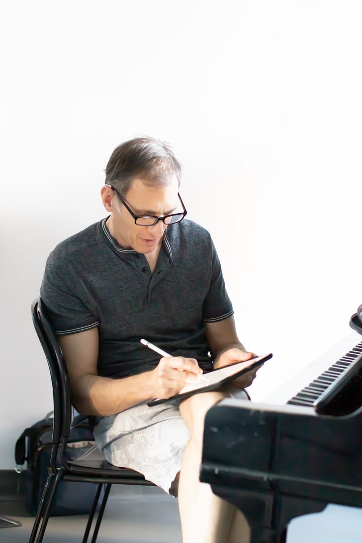 Susan Derry - woman in profile, medium length brown bob and black sweater, hand on chin. editing the script in a rehearsal room. Howard Breitbart making notes on an iPad while sitting at a piano.