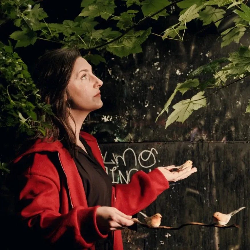 A photograph of poet Bridie Squires standing beneath leafy branches outdoors, holding out her hands as small birds perch on her fingers and nearby twigs. She wears a red jacket and looks upward toward the foliage, with soft natural light filtering through the leaves, creating a calm, reflective atmosphere.